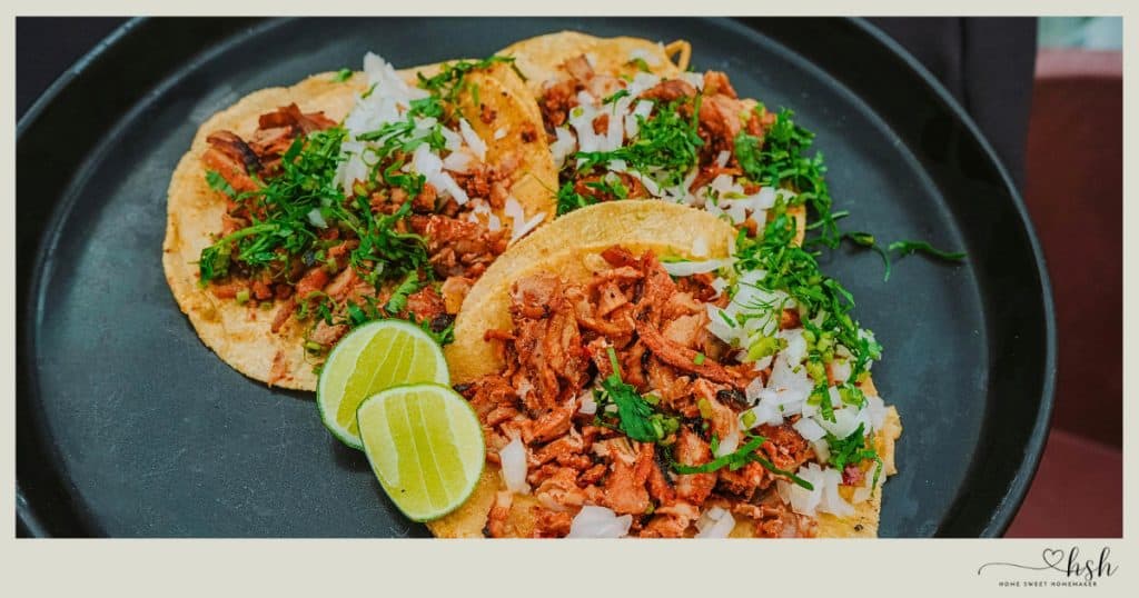 Street-style tacos with seasoned meat, fresh cilantro, diced onions, and lime wedges on dark plate showing quick winter meal prep dinner idea for busy moms.